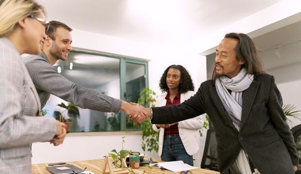 Two men shaking hands around a desk while two women on either side look on