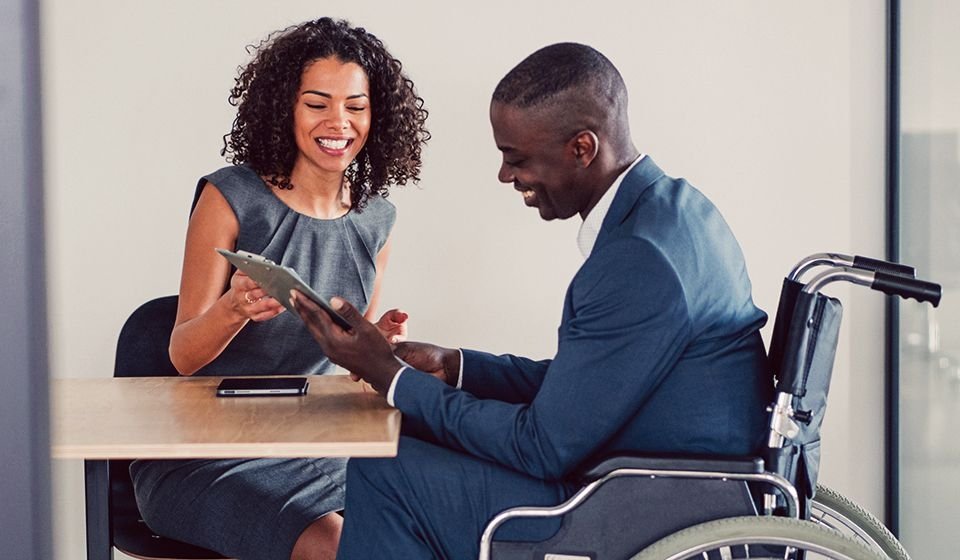 Two people, a woman and a man, sit at a desk looking at a tablet